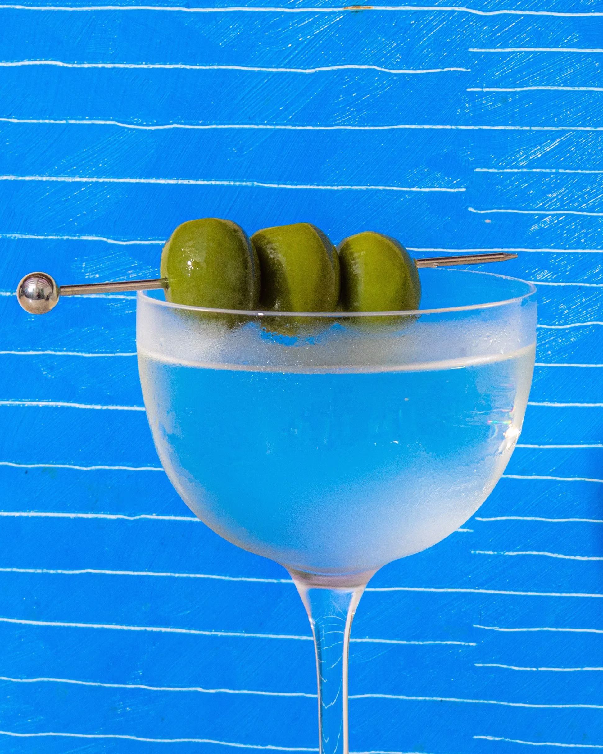 Chilled martini with three green olives on a skewer, blue background.