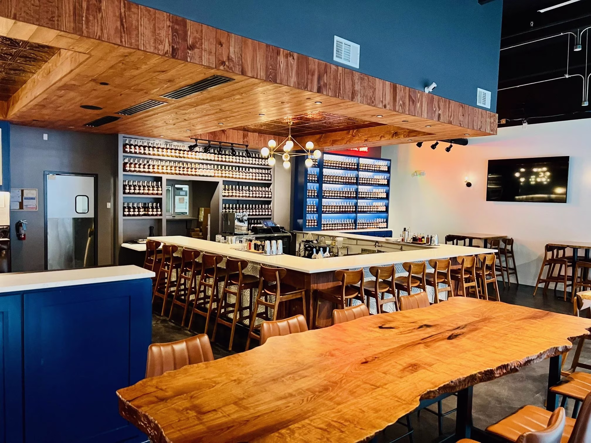 Modern bar interior with long live-edge table and extensive bottle displays.
