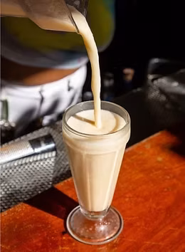 Thick, light-colored drink being poured into a tall, ribbed glass.