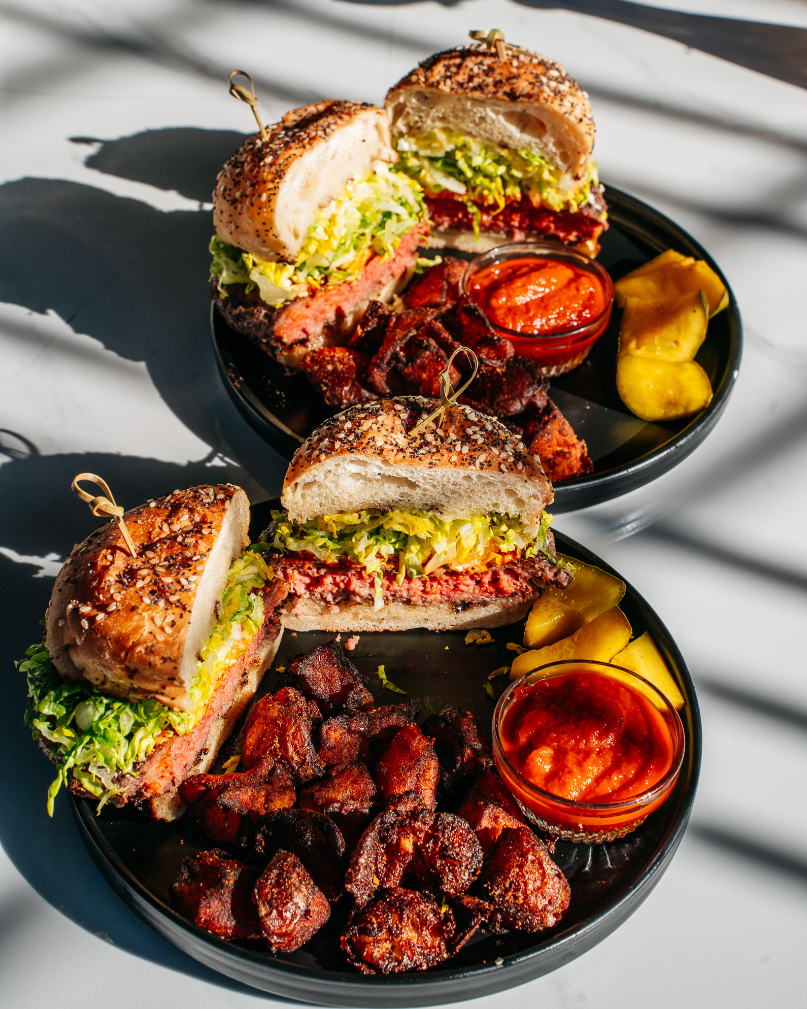 Two plates with sliced burgers, roasted vegetables, sauce, and pickles in sunlight.