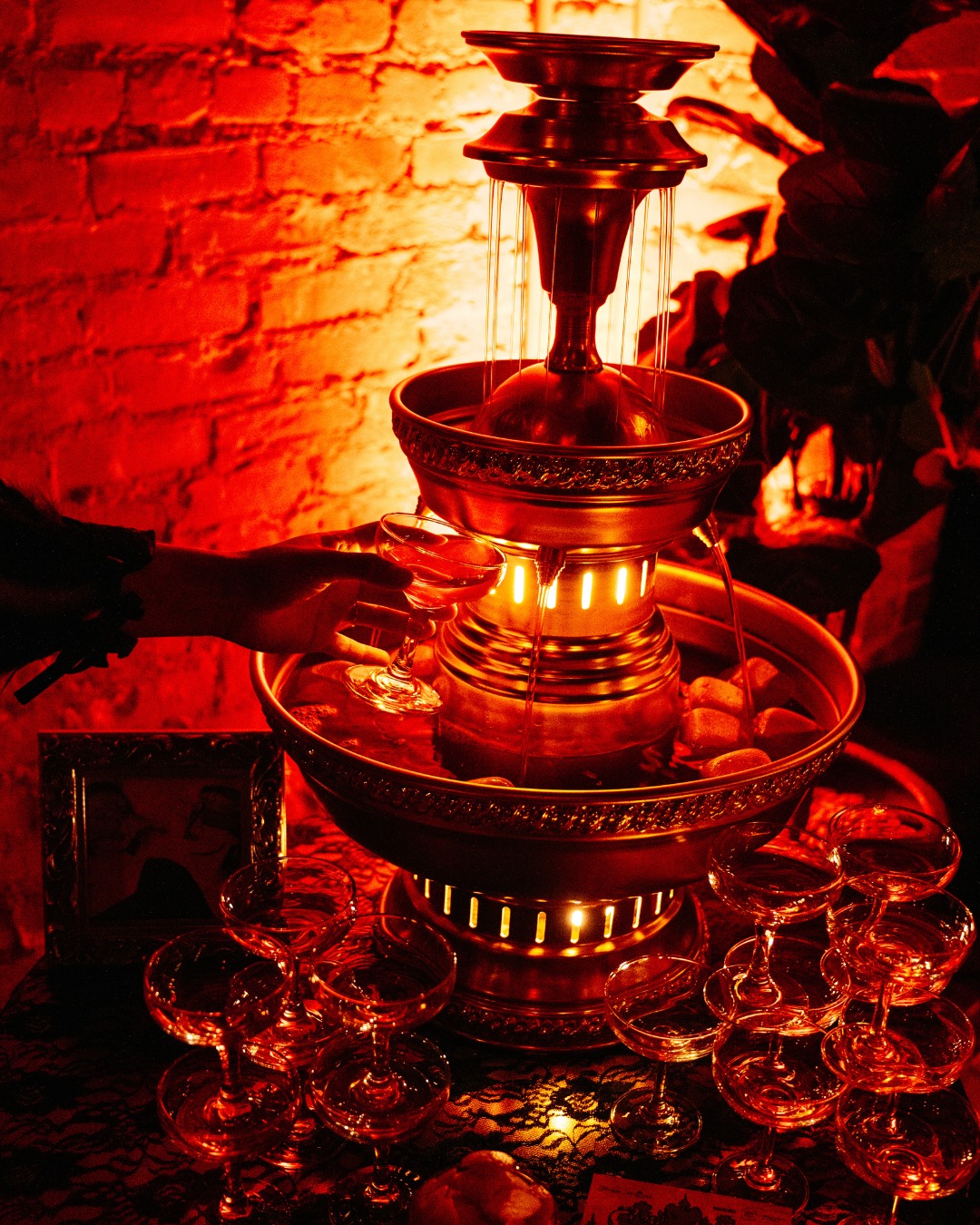 Hand fills coupe glass from a red-lit tiered drink fountain.