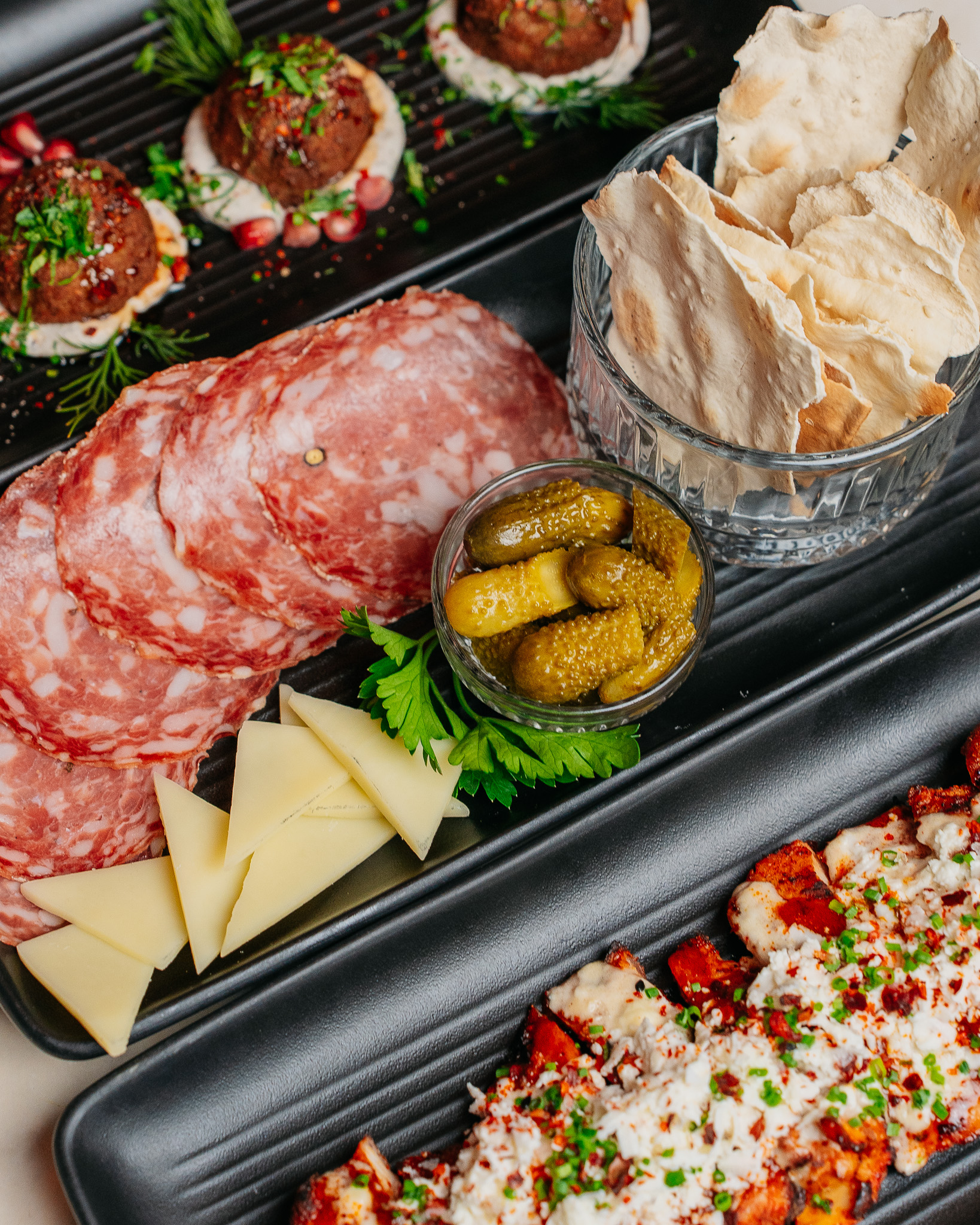 Salami, cheese, pickles, flatbread, and various prepared dishes on dark trays.