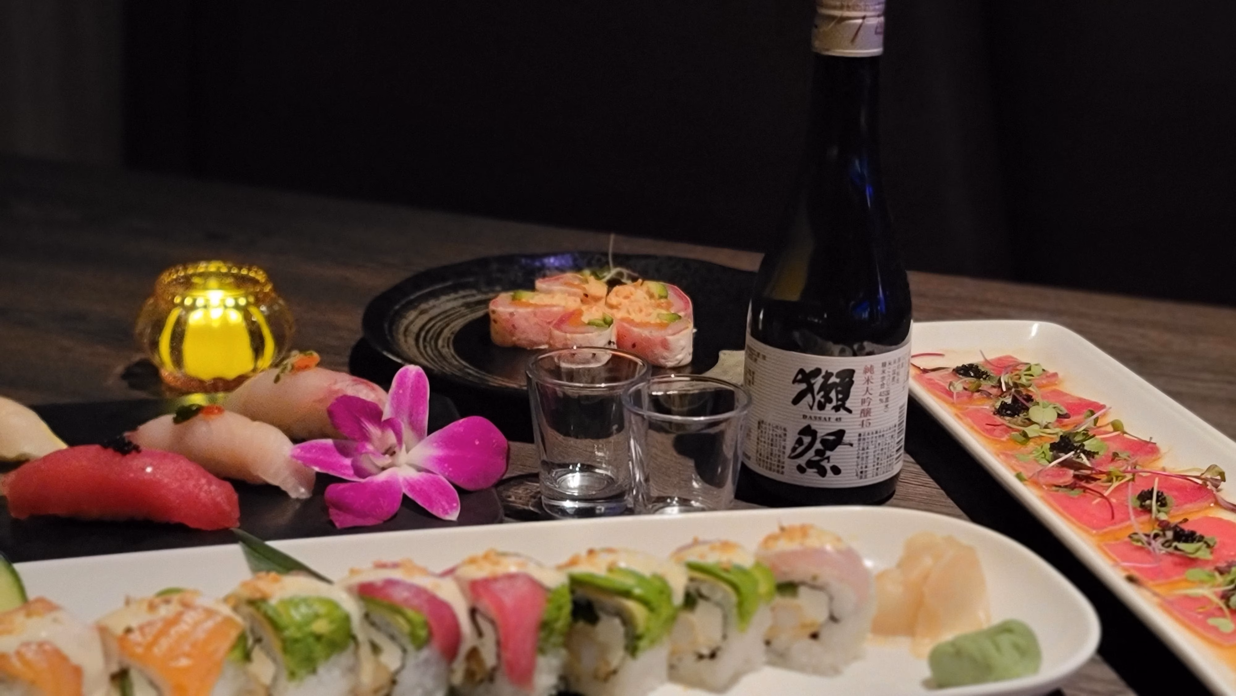 Assortment of sushi, sashimi, and sake on a dark table with an orchid.
