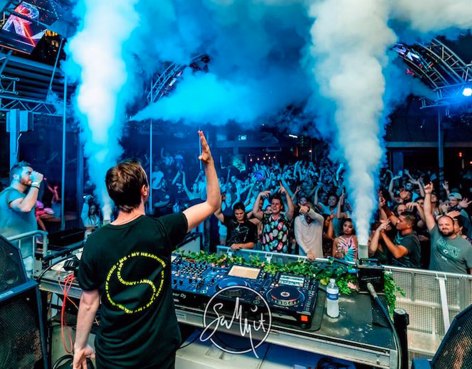 DJ with raised arm facing crowd under blue lights and smoke.