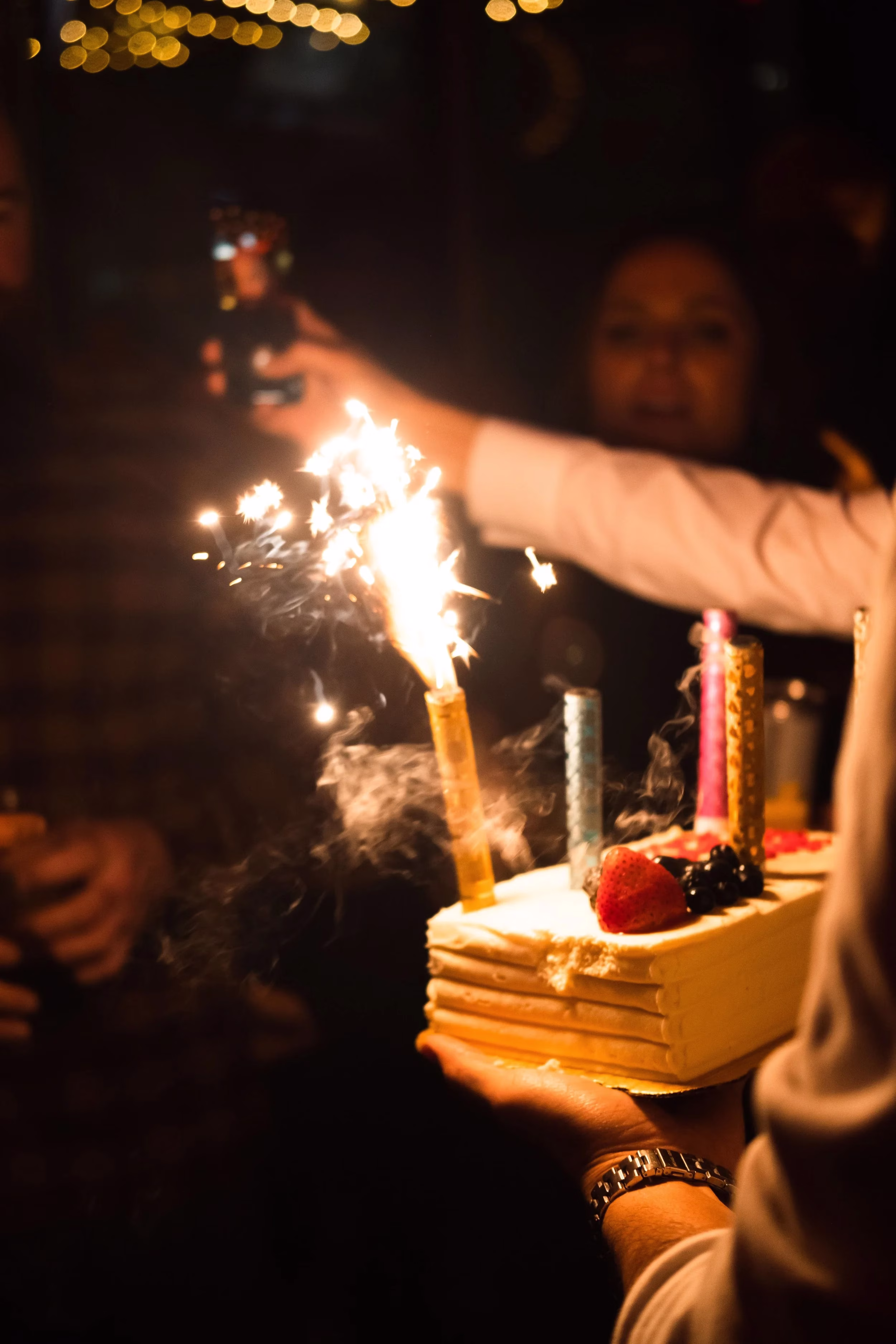 Person holding a cake with a lit sparkler, candles, and fruit.