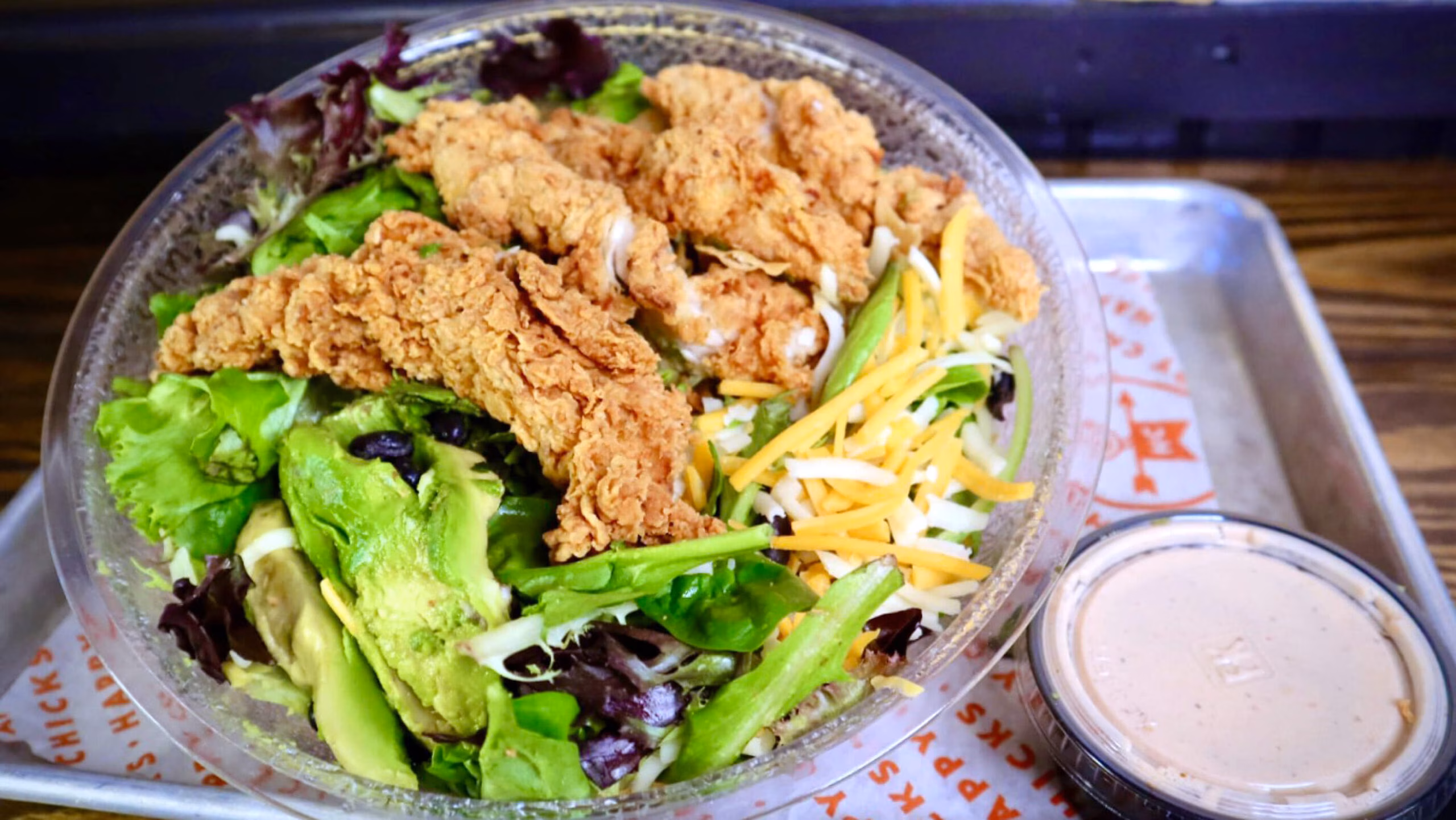 Fried chicken salad with avocado, cheese, and dressing in a plastic bowl.