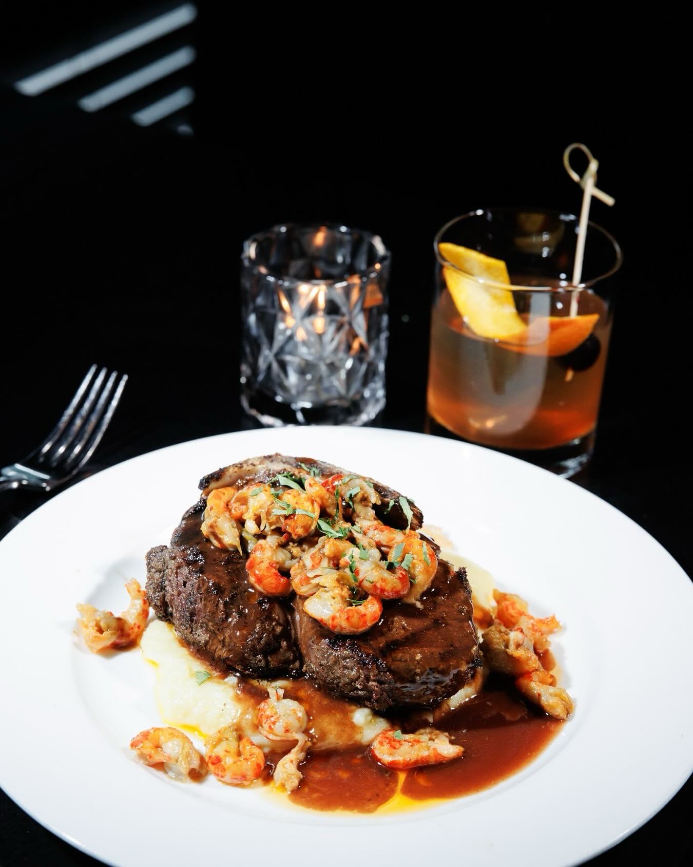 Steak and shrimp on mashed potatoes with sauce, cocktail, and candle.