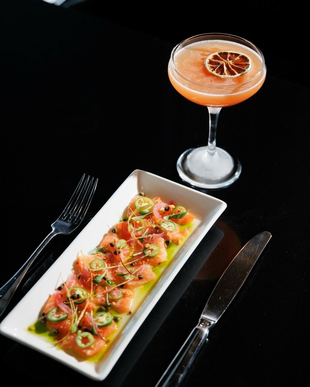 Thinly sliced salmon, cocktail, fork, and knife on a black table.