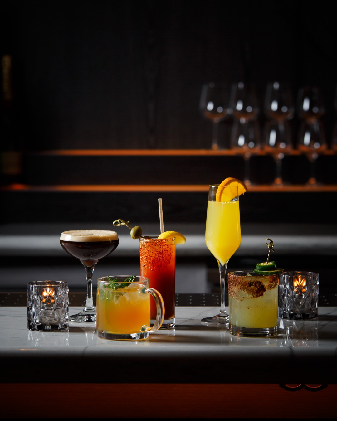 Five colorful cocktails and candles on a reflective bar top.