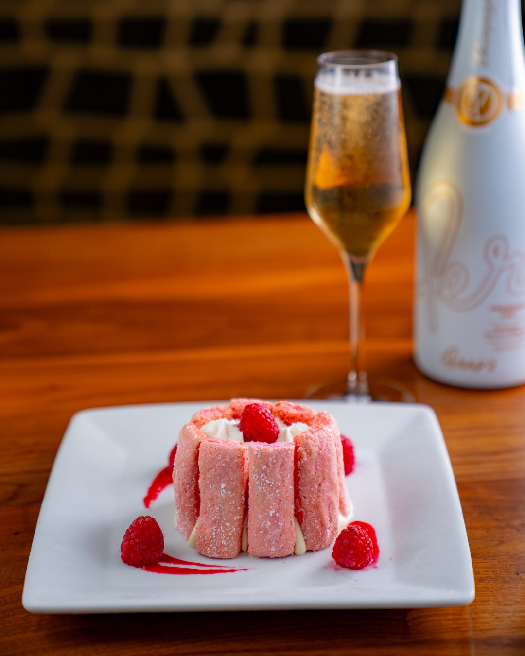 Pink dessert with raspberries, sparkling wine glass, and bottle on table.
