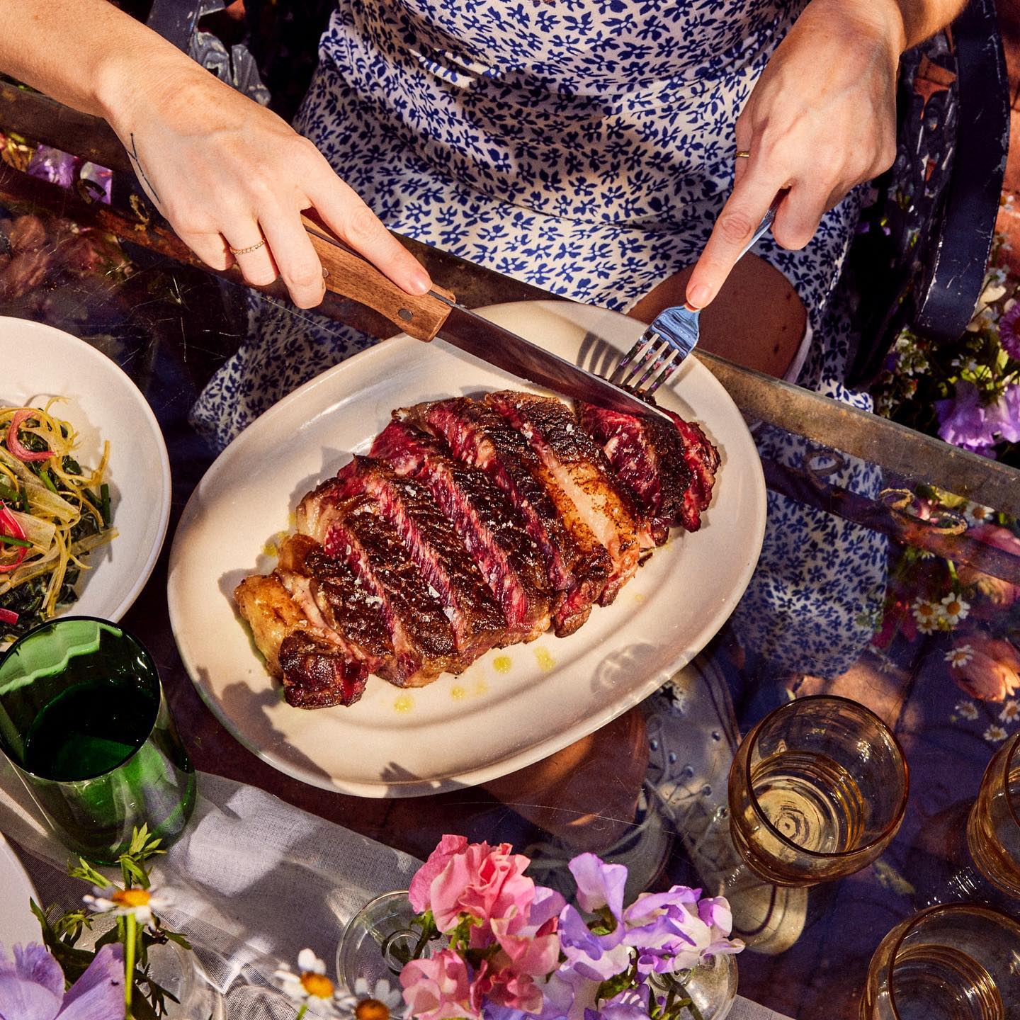 Hands cutting a large, sliced steak on a plate outdoors.