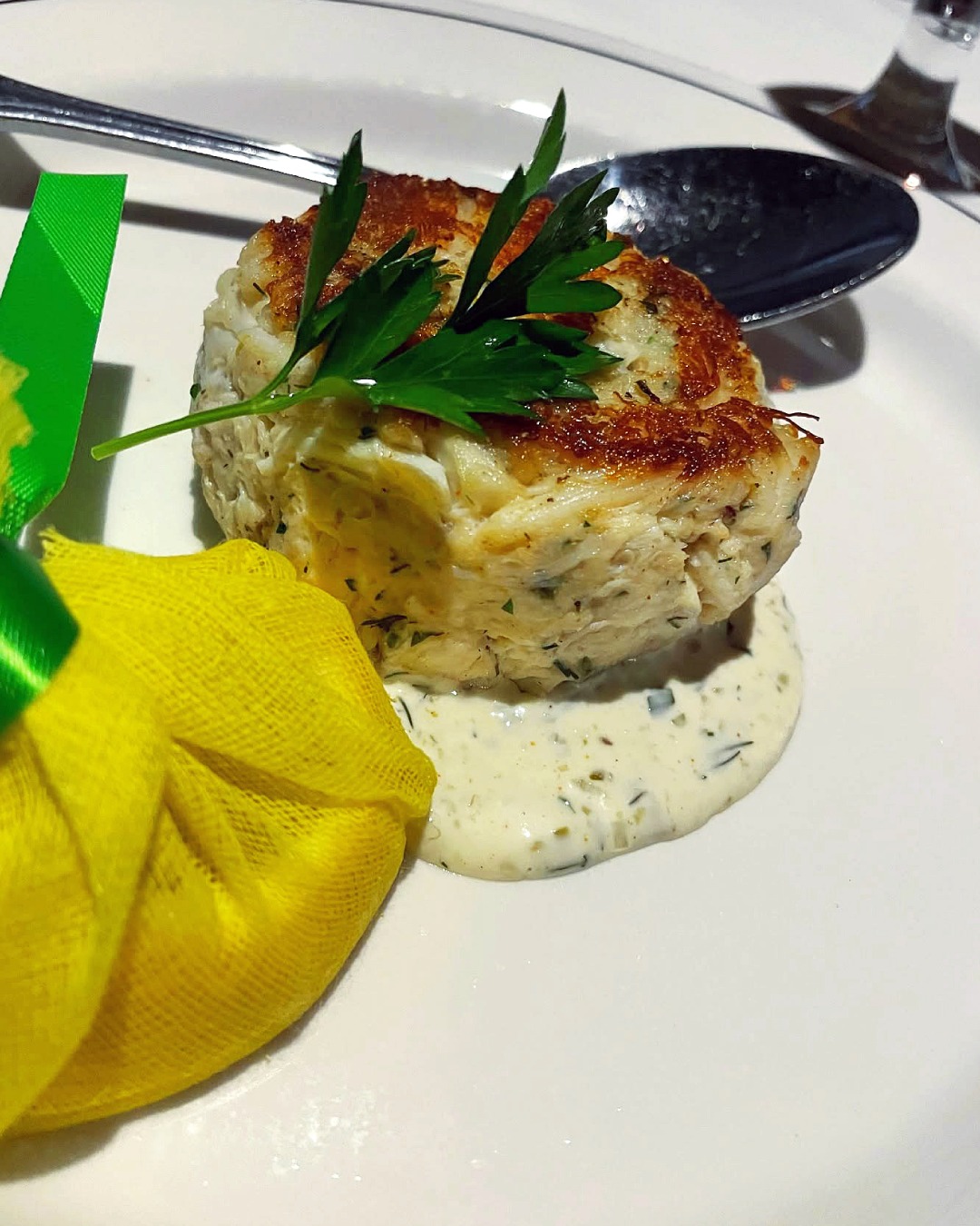 Crab cake with white sauce, parsley, and lemon in mesh bag.