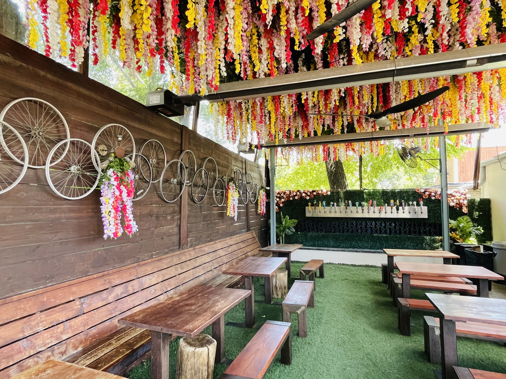 Outdoor patio with colorful hanging flowers, bicycle wheel decor, and wooden tables.
