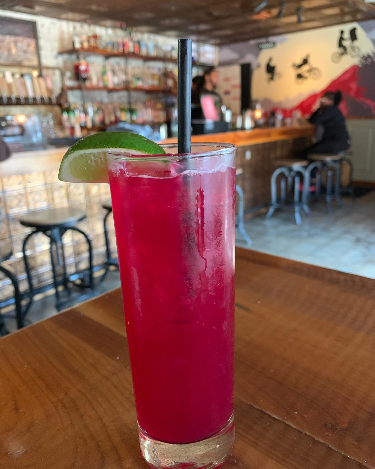 Red drink with lime and straw on a wooden table at a bar.