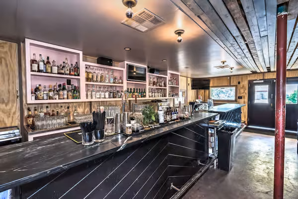 Interior of a bar with black counter, pink shelves, and wooden ceiling.