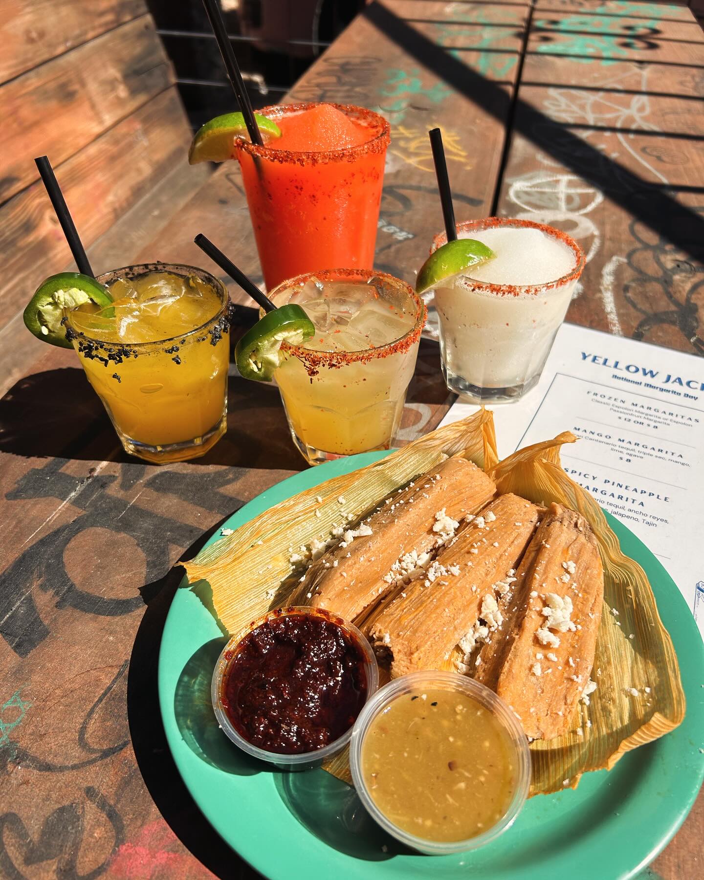 Four margaritas and a plate of tamales with sauces on a wooden table.