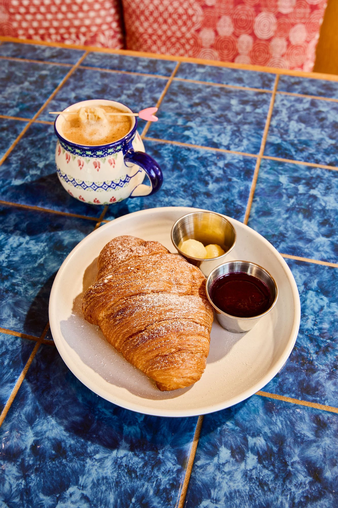 Croissant, jam, butter, and coffee on a blue tiled table.