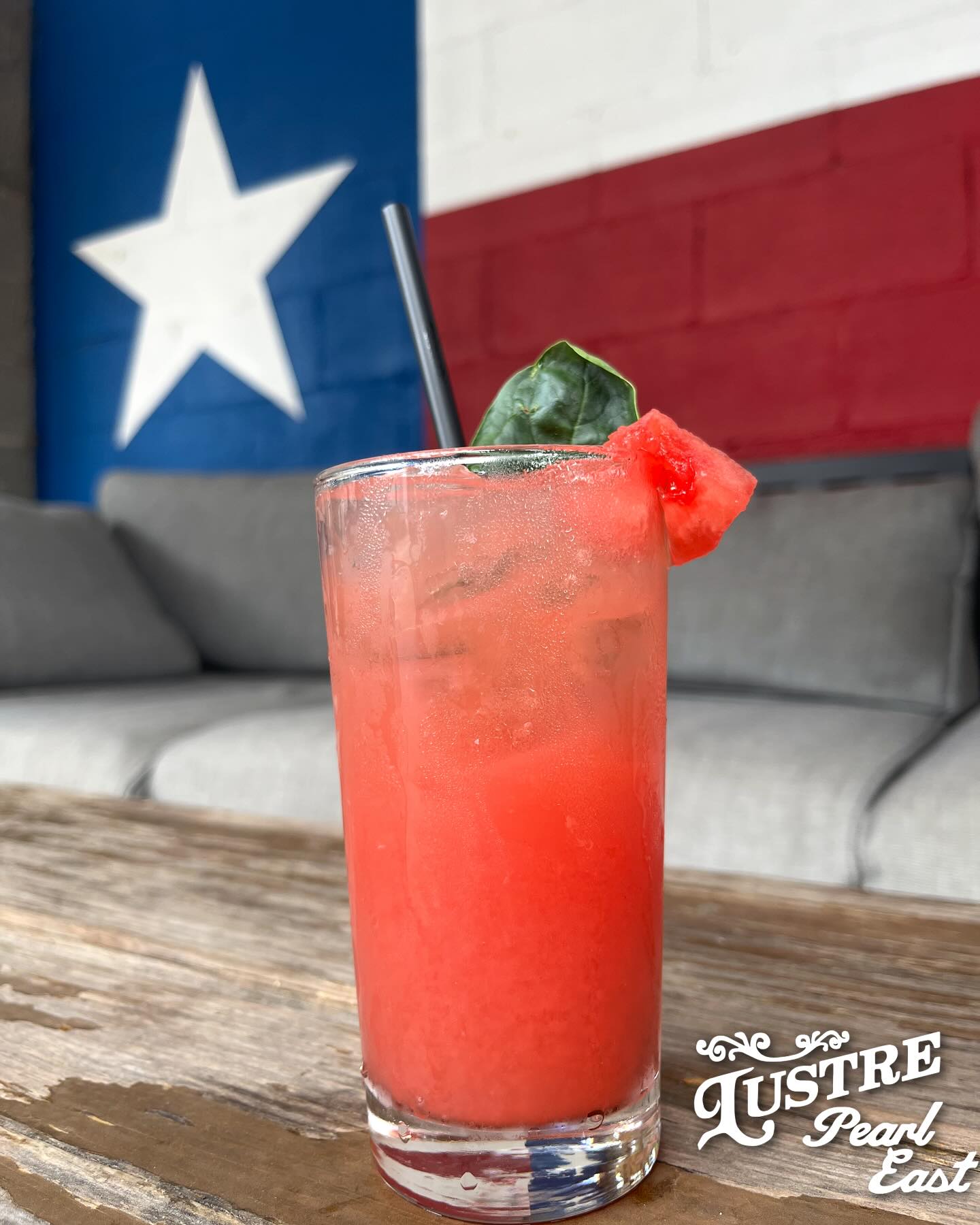 Watermelon-garnished red drink on wooden table, Texas flag background.