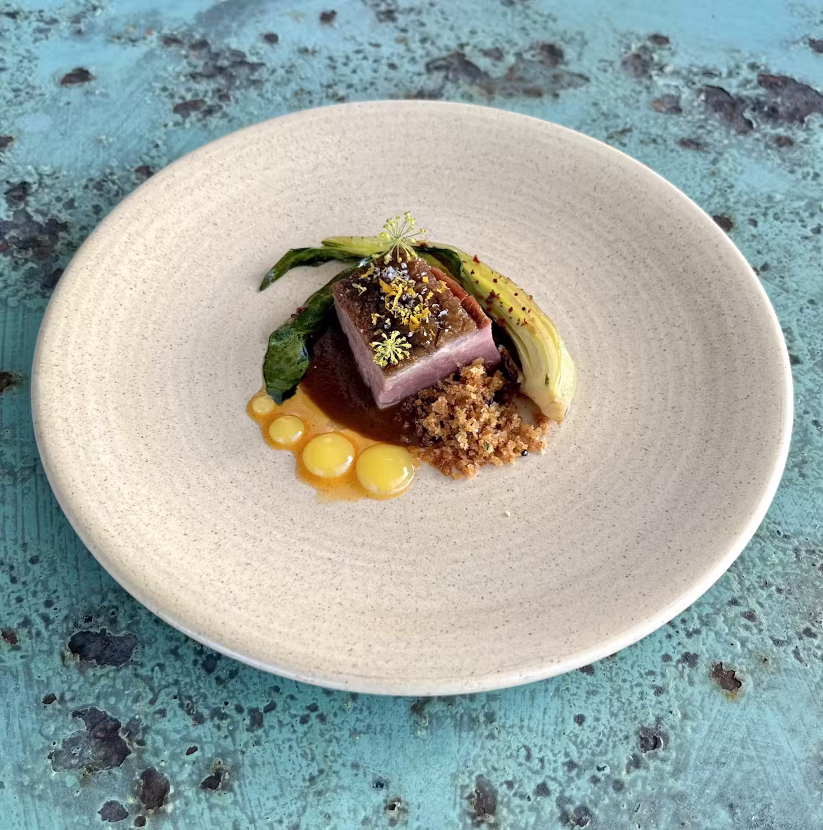Plated dish with meat, sauces, green leaf, and crunchy topping.