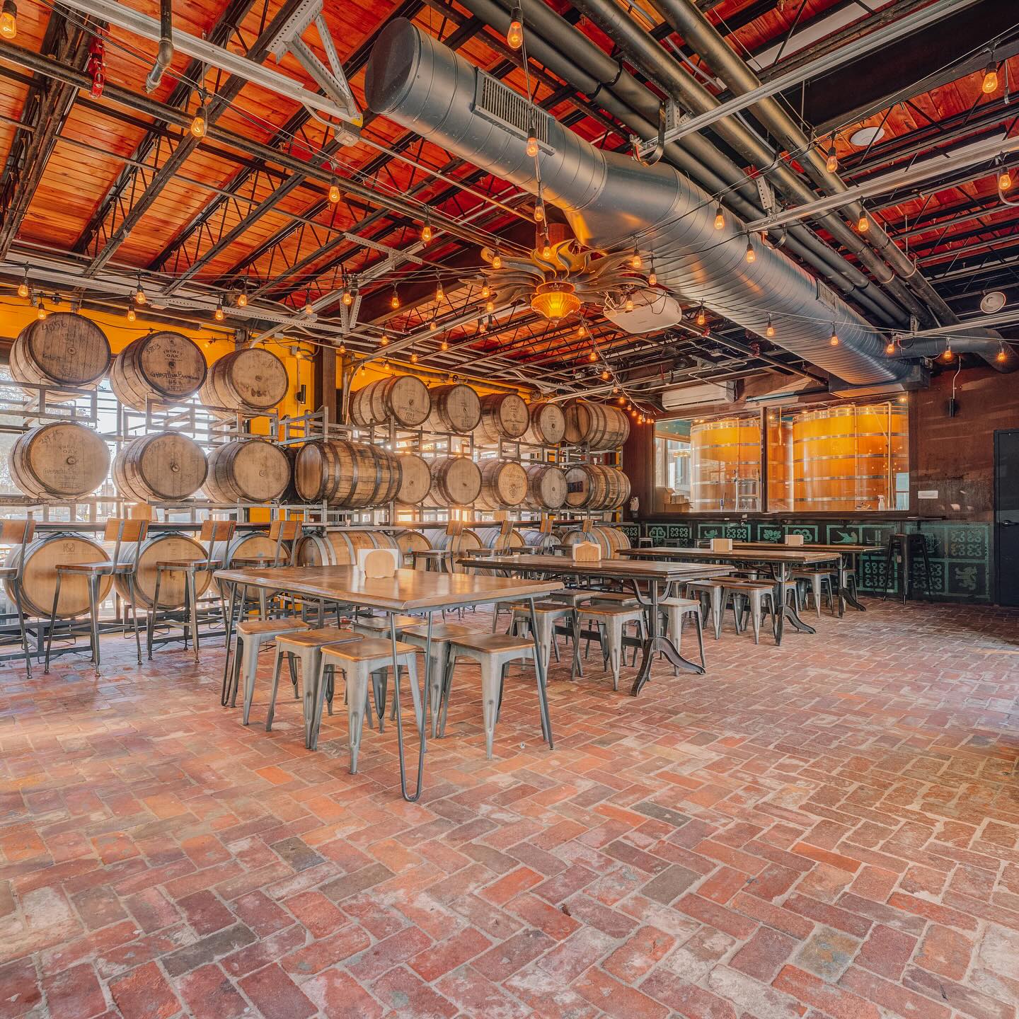 Rustic room with stacked wooden barrels, brick floor, tables, and ceiling ducts.