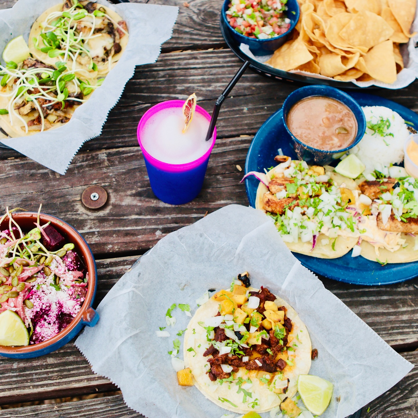 Various tacos, chips, salsa, and frozen drink on a wooden table.