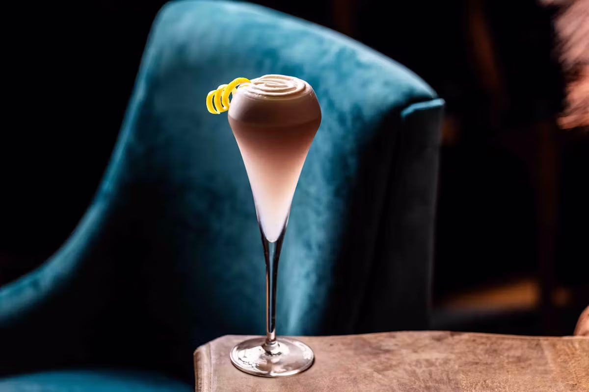 Layered cocktail with foam, lemon spiral, against a velvet chair.