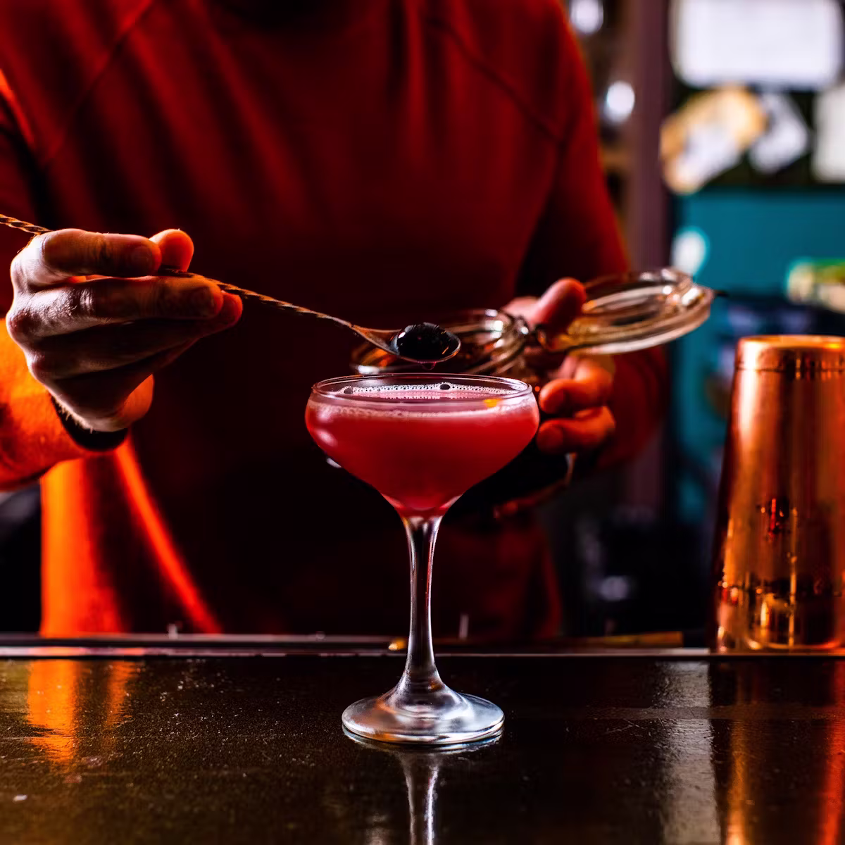 Hands garnishing a red cocktail with a dark berry.