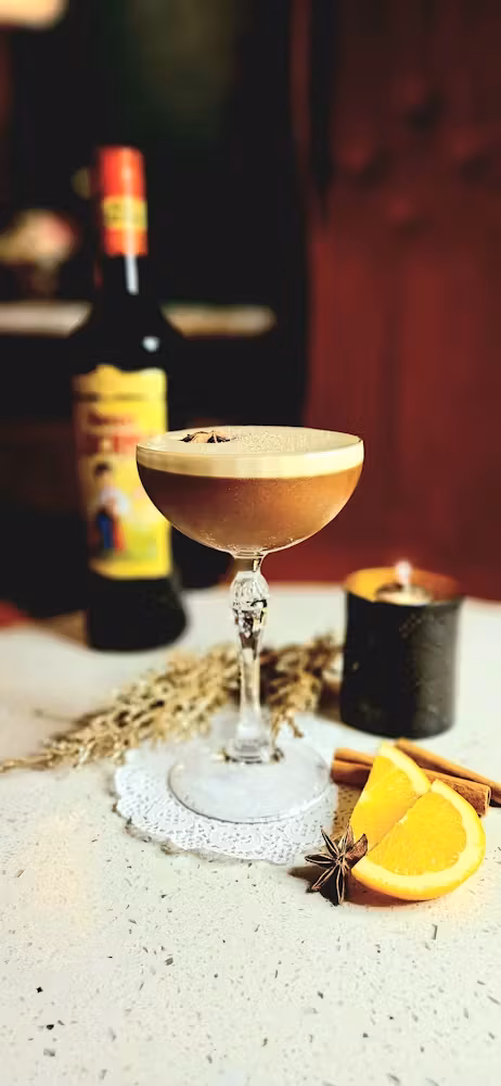 Brown cocktail with foam, star anise, orange, cinnamon, and a bottle.