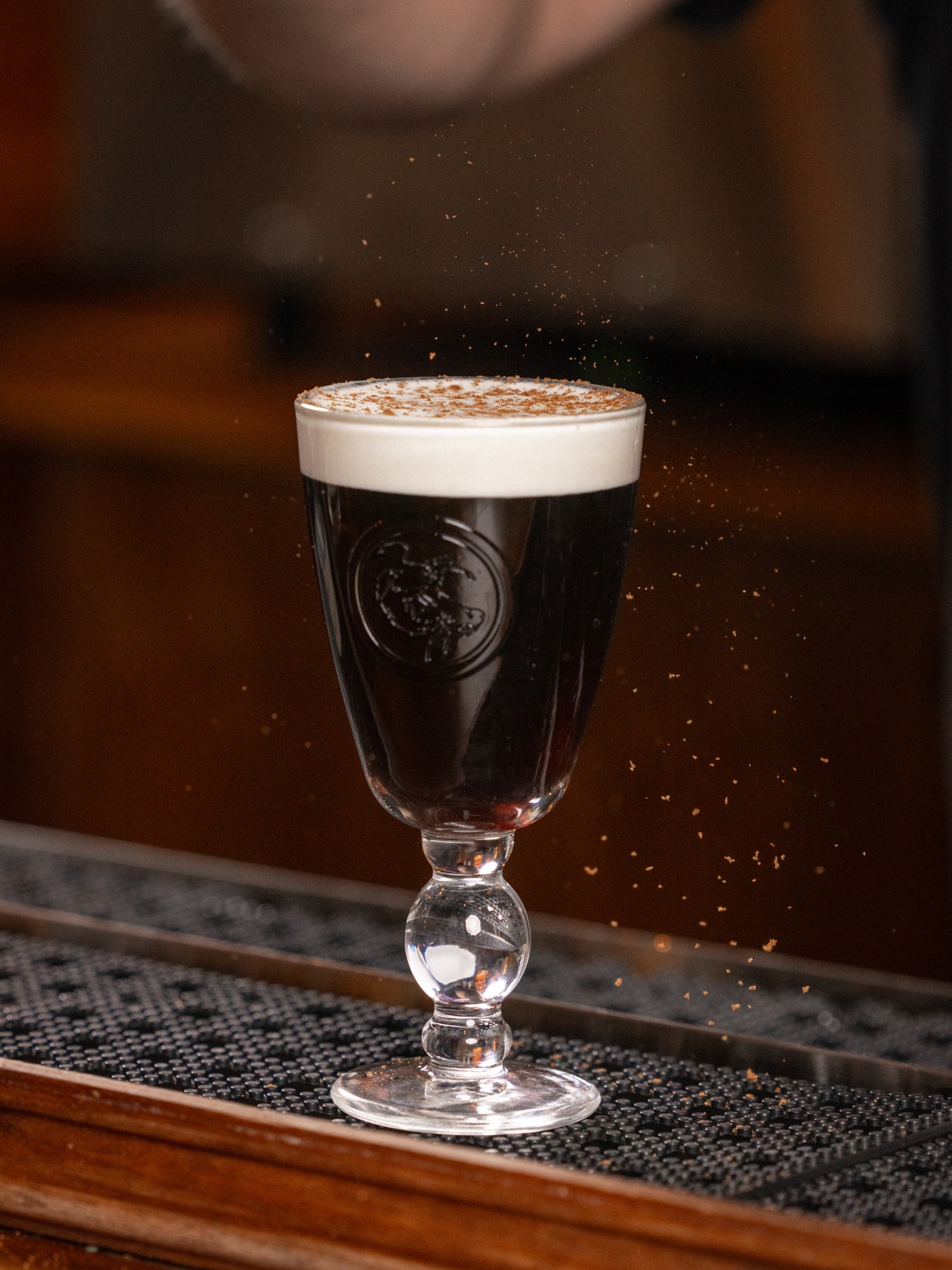 Dark beverage in stemmed glass with white foam and falling brown sprinkles.