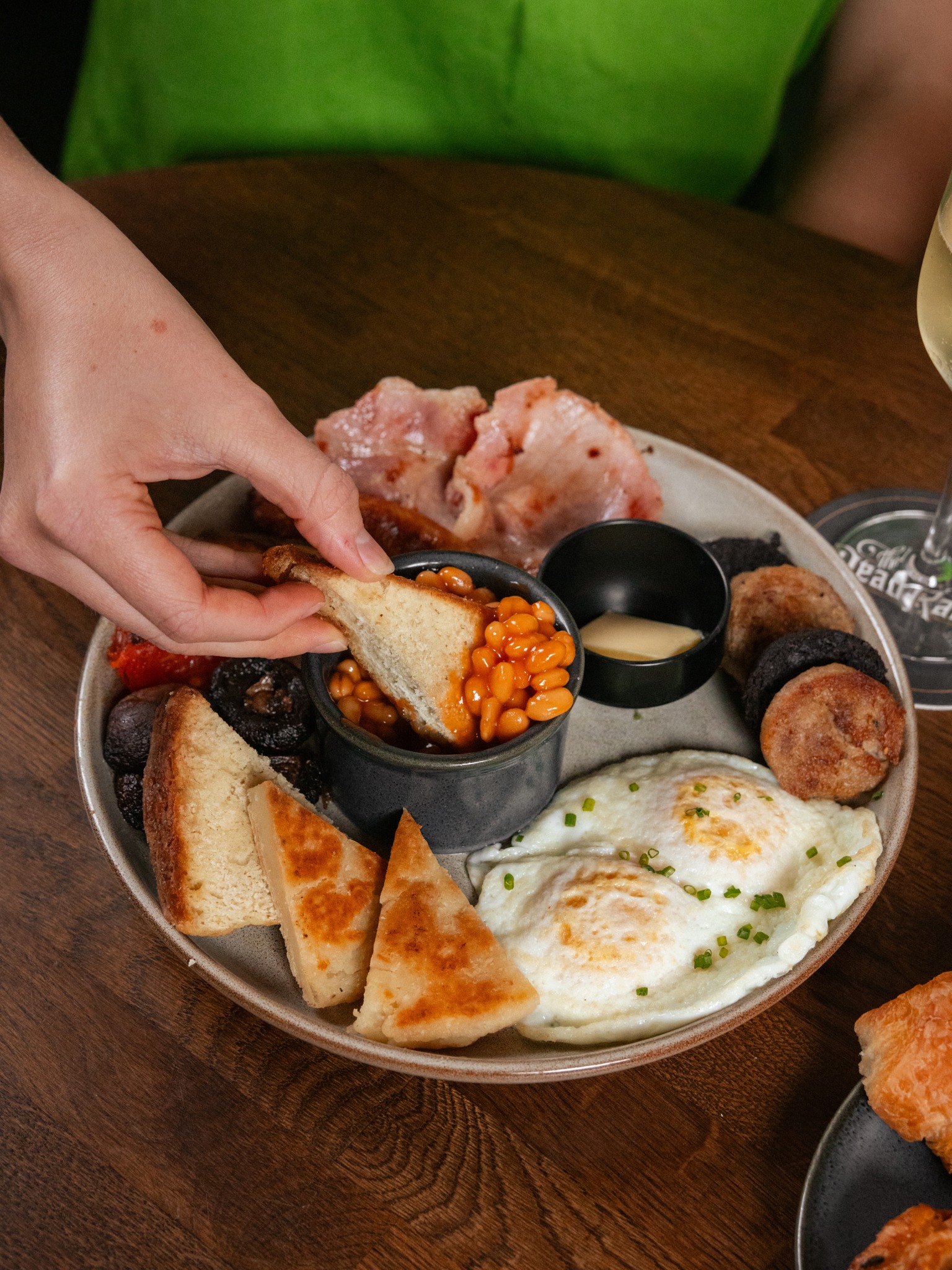 Hand dipping toast into baked beans on a full English breakfast plate.