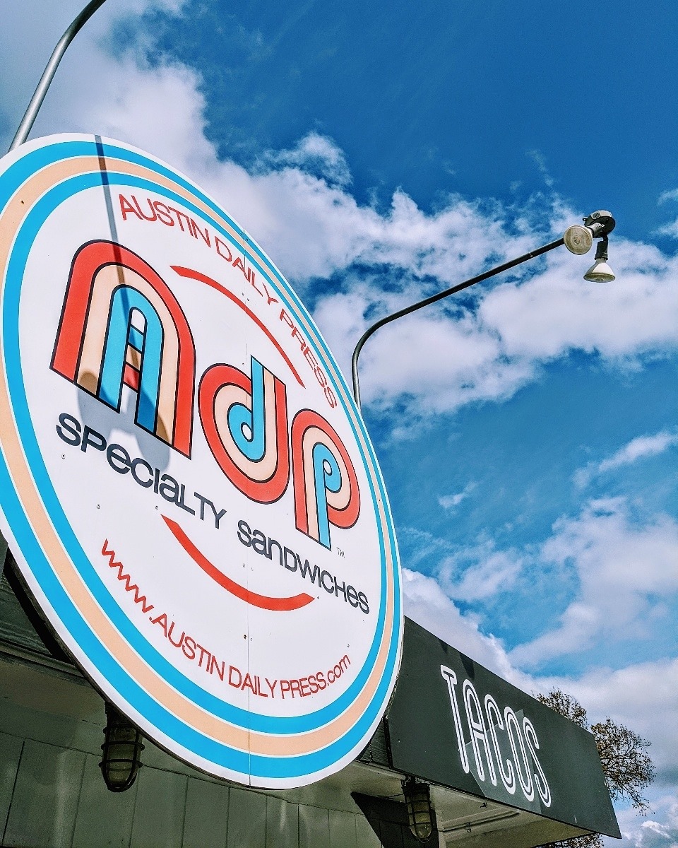 Round Austin Daily Press sign and part of a "Tacos" sign under blue sky.