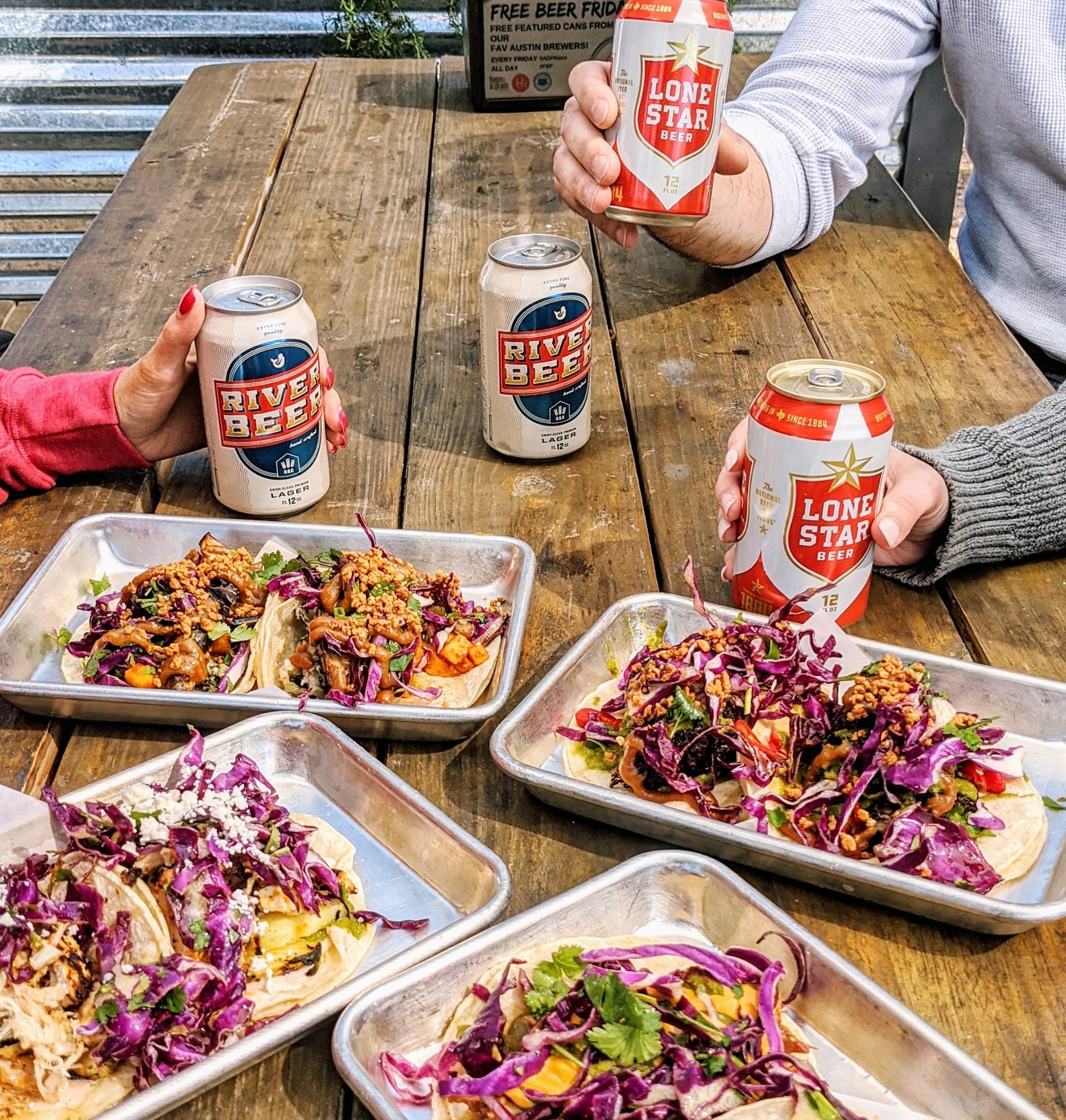 Plates of colorful tacos and beer cans on a wooden table.
