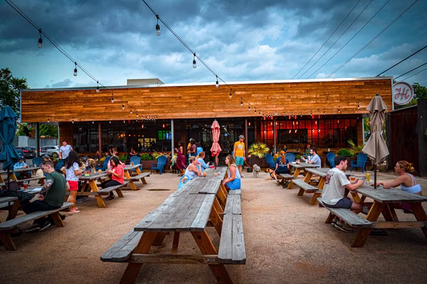 People gather at an outdoor patio with picnic tables and string lights.
