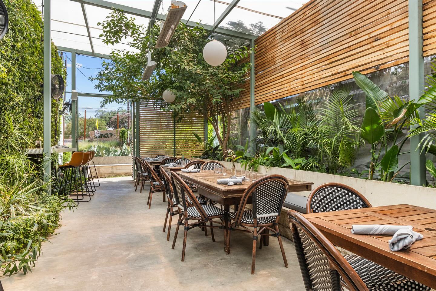 Covered outdoor restaurant patio with tables, chairs, and abundant greenery.