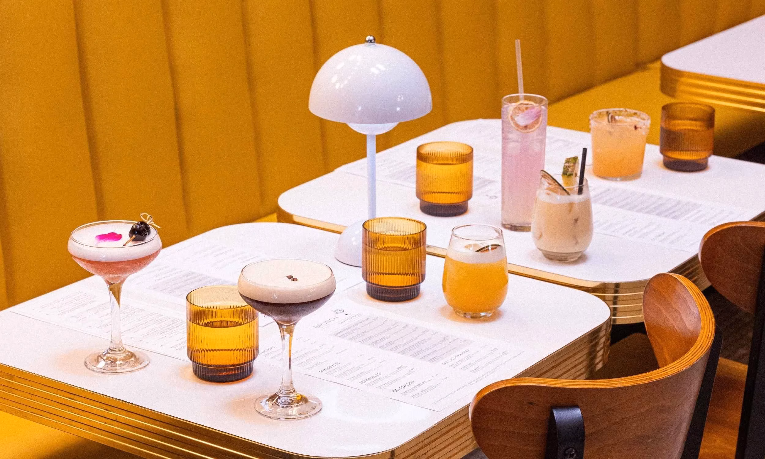 Diverse cocktails and drinks on white tables against a yellow background.