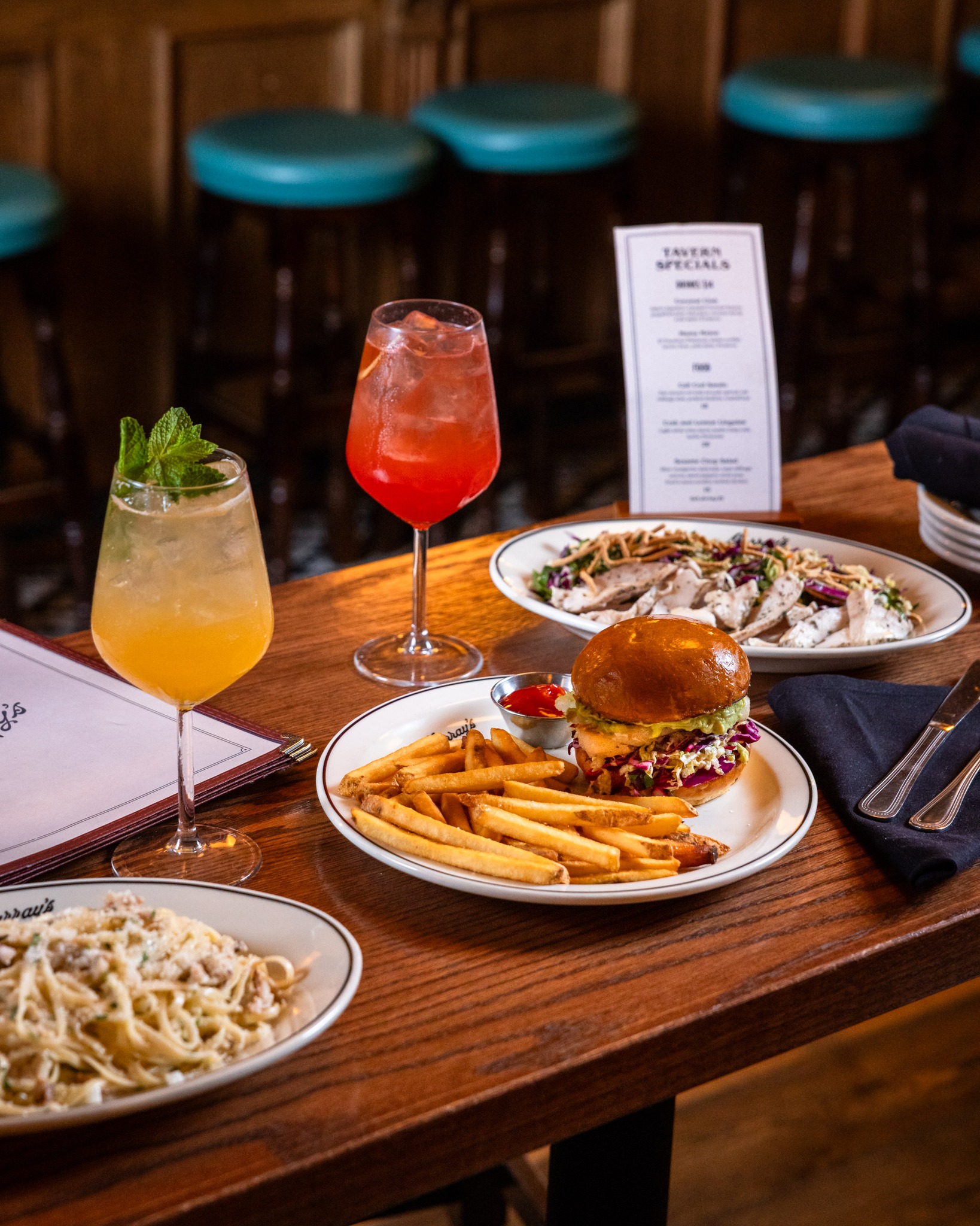 Burger, fries, pasta, salad, and two cocktails on a wooden table.
