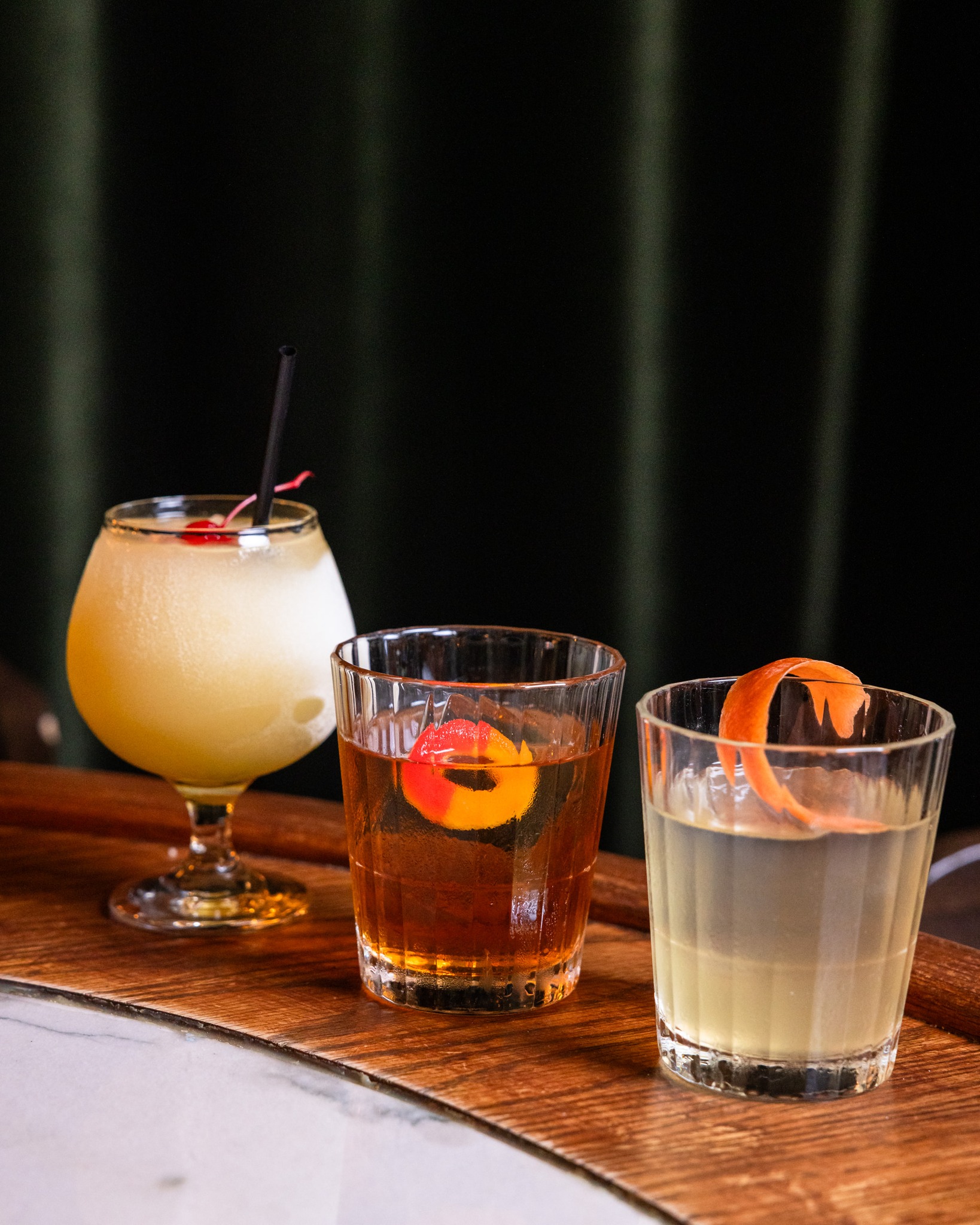 Three cocktails: frozen, amber, and light, on a wooden tray.