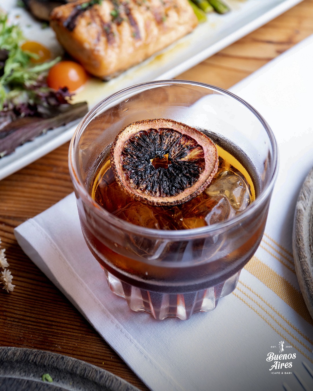 Glass of drink with dried orange, ice, and blurred grilled salmon.