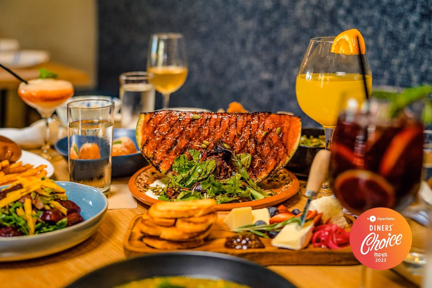 Restaurant table with grilled watermelon, cheese board, salads, and cocktails.