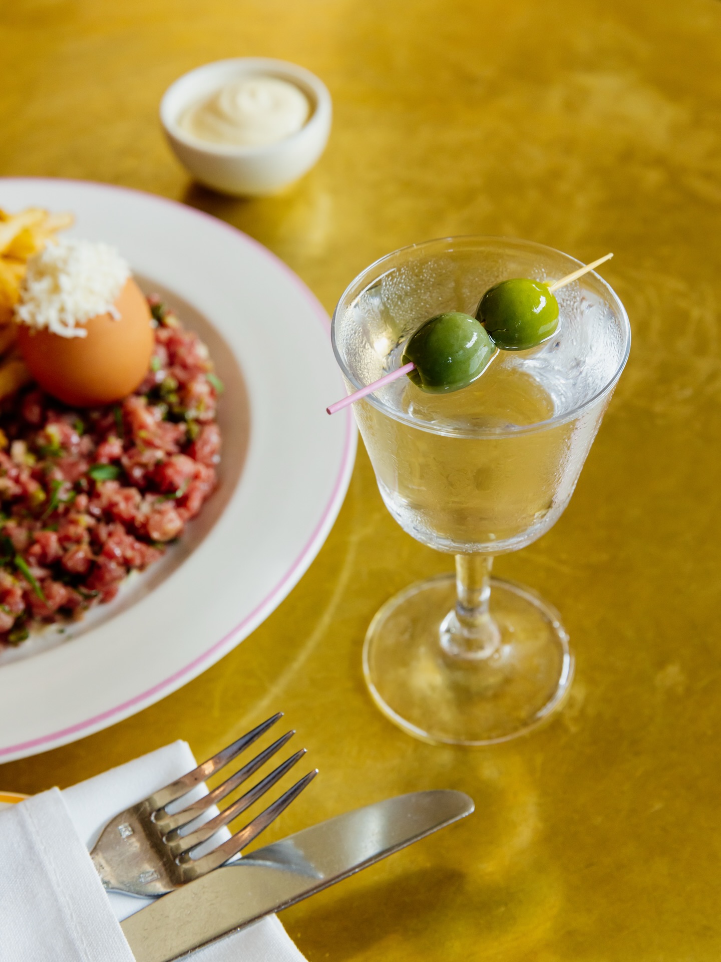 A martini, steak tartare with egg, and fries on a gold table.