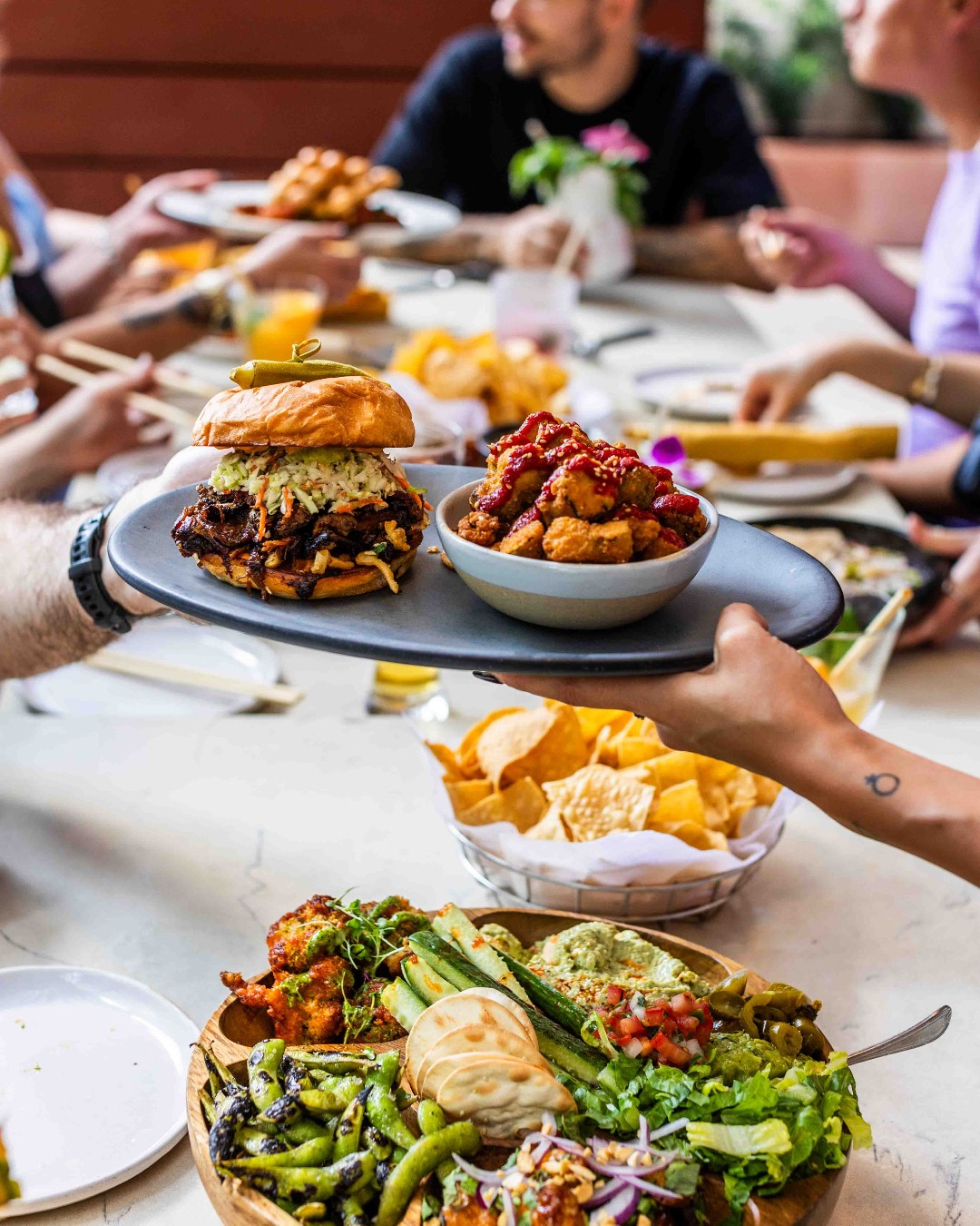 Hands serving a burger and chicken bites alongside other food.