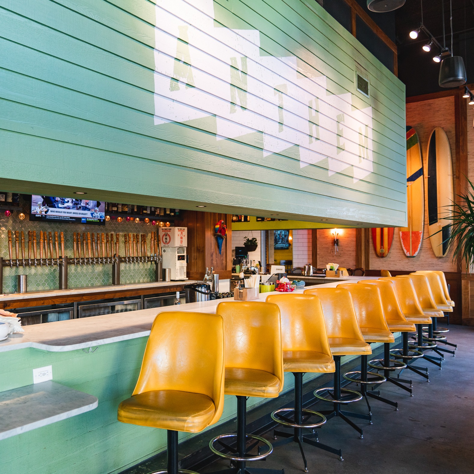 Bar interior with yellow stools, beer taps, a TV, and surfboards.