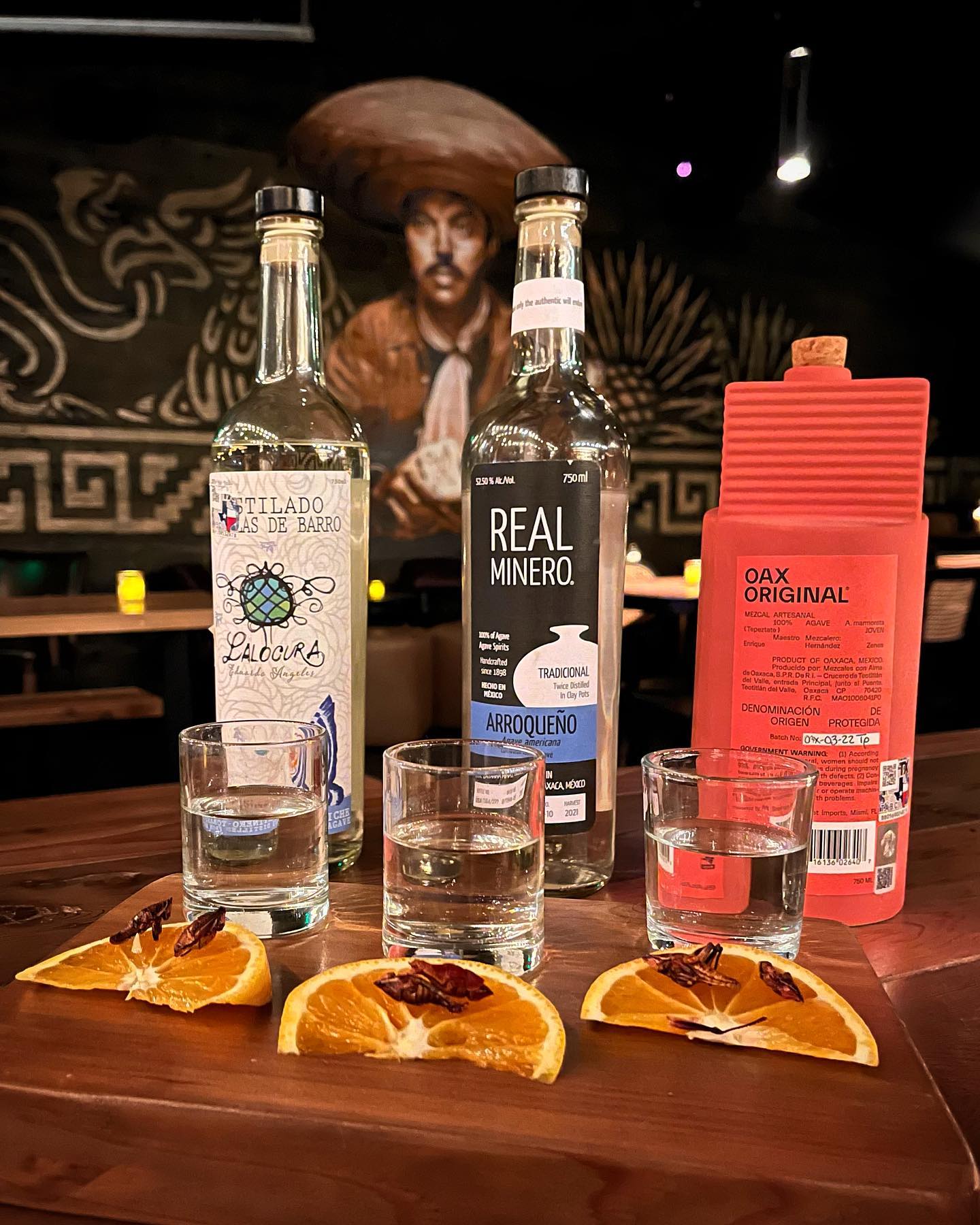 Three mezcal bottles, shots, and orange slices with chapulines.
