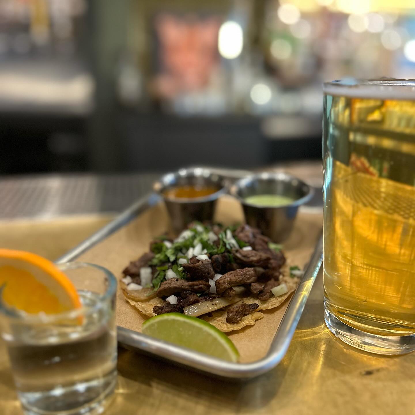 Tacos, beer, shot, sauces, lime, and orange on a tray.