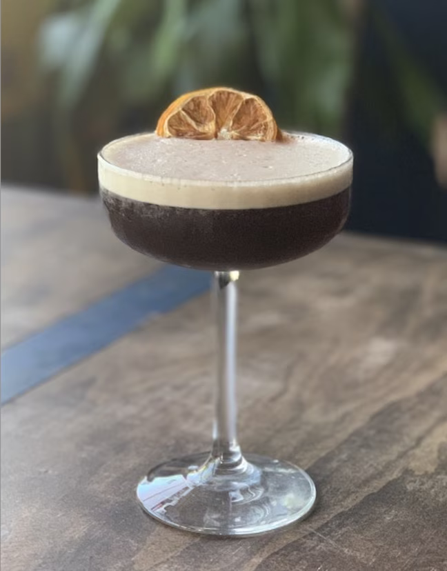 Dark cocktail in coupe glass with foam and dried orange garnish.