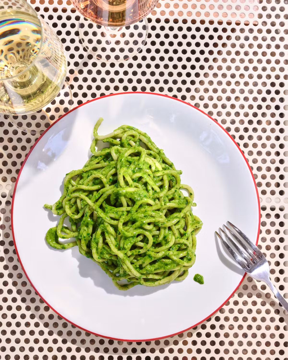 Green pasta on a white plate with fork and wine glasses.
