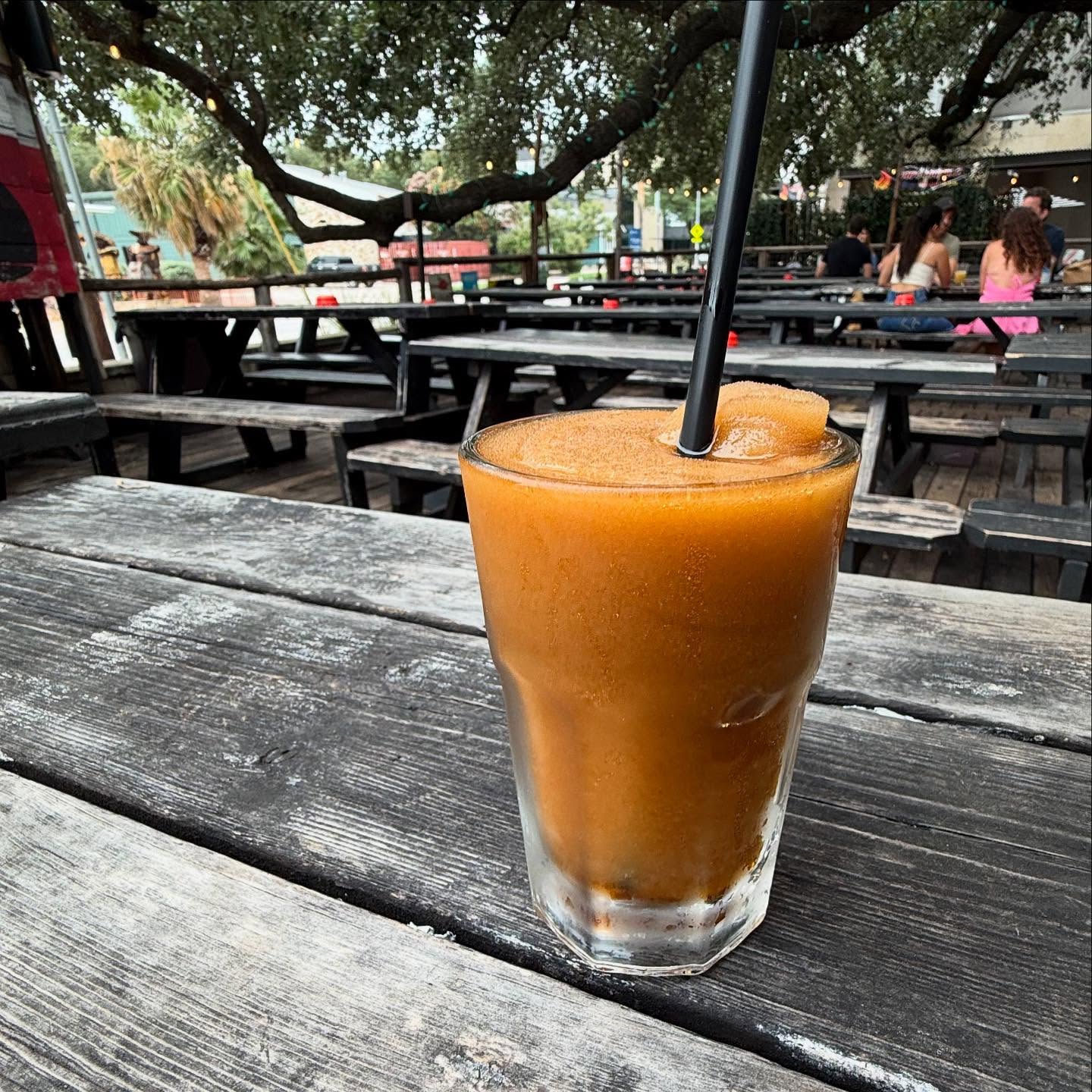 Glass of frozen orange drink with straw on a weathered picnic table.