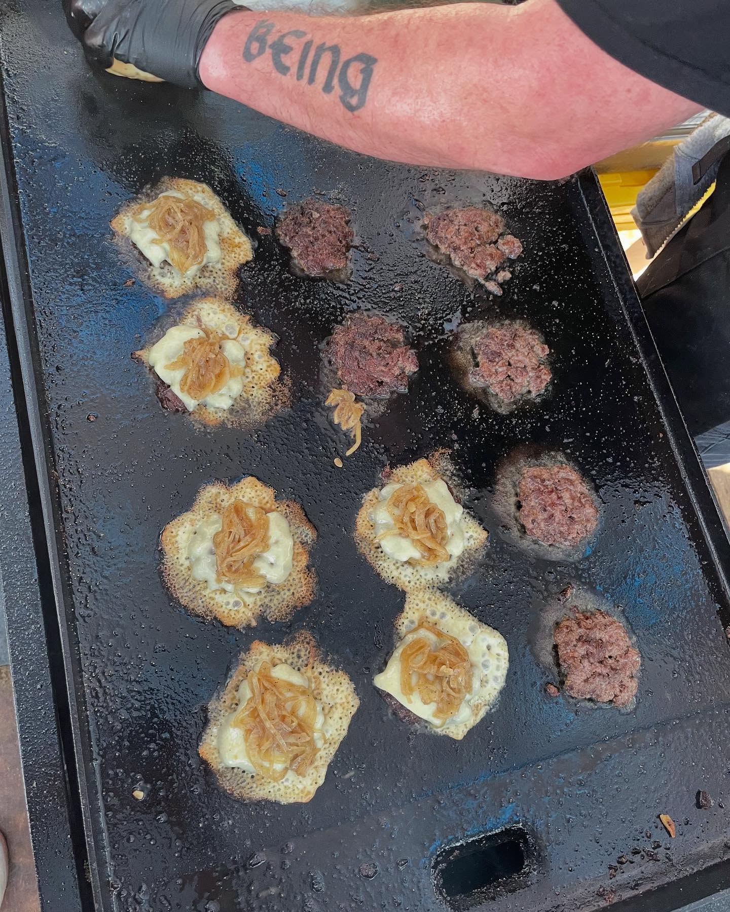 Burgers with crispy cheese, caramelized onions cooking on a griddle.