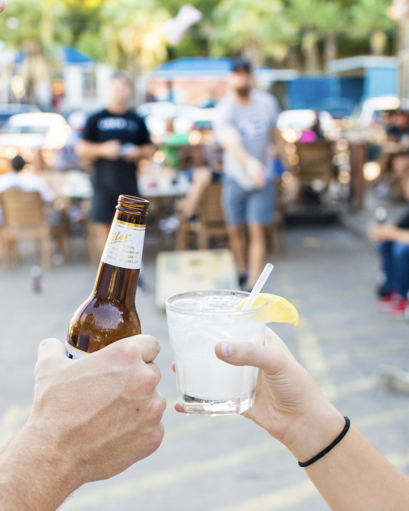 Hands toasting a beer bottle and a lemon drink outdoors.