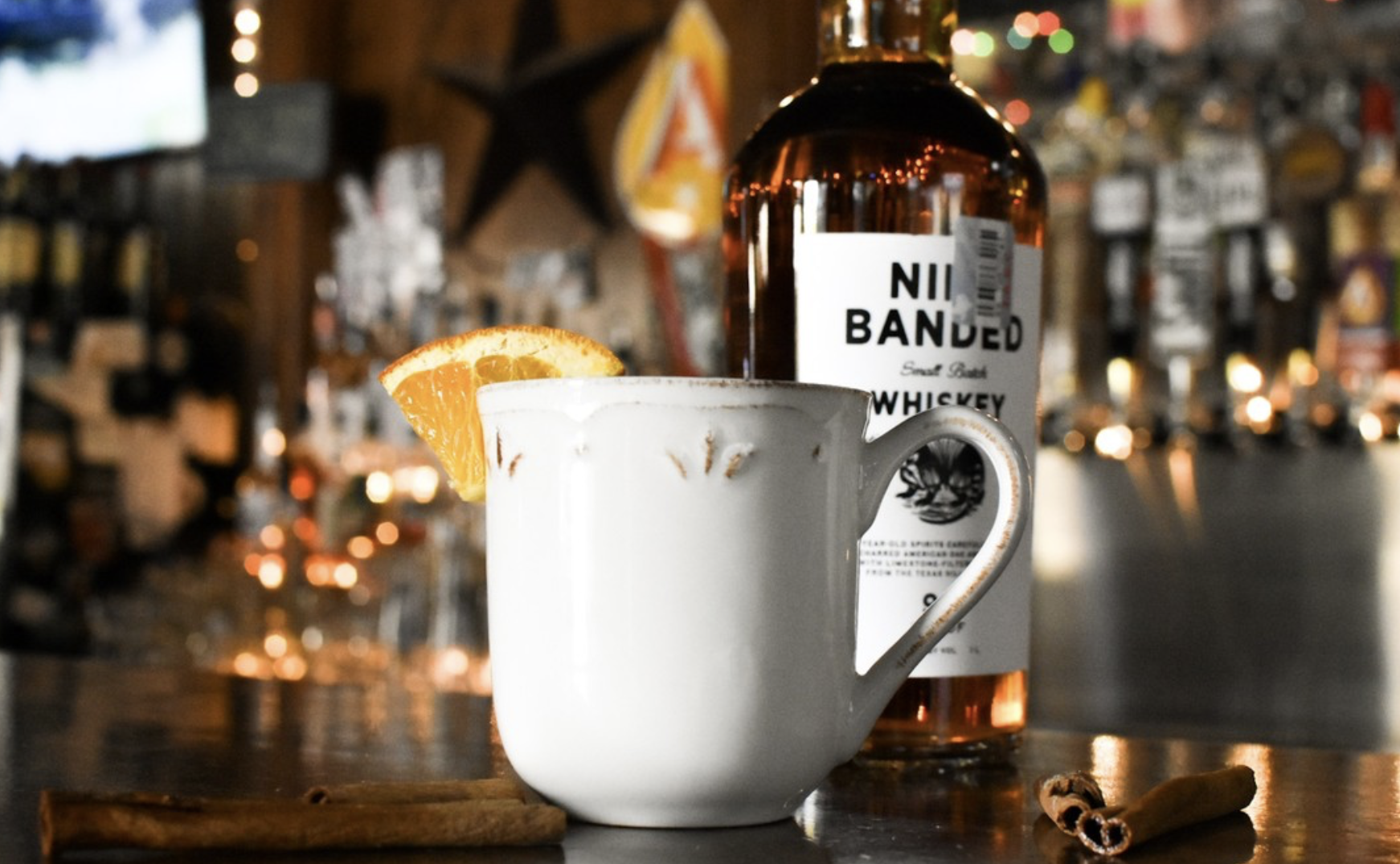 White mug with orange slice, whiskey bottle, and cinnamon sticks on a bar.